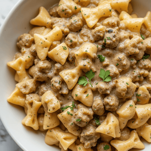 A bowl of creamy beef and bowtie pasta, richly coated in a velvety cream sauce with bits of ground beef and garnished with fresh parsley, served on a clean white plate with bright, natural lighting for a homemade and inviting look.